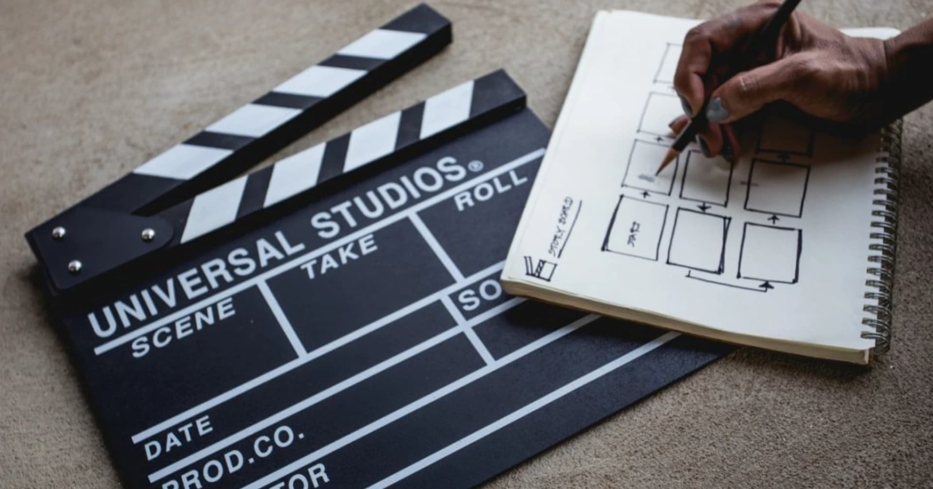 Foto de uma claquete de cinema da Universal Studios ao lado de um caderno espiral onde uma mão desenha um storyboard, como metáfora de preparar situações em vez de roteiros.