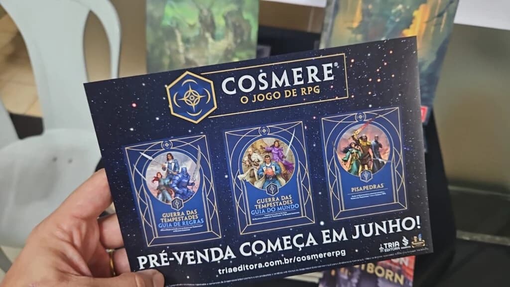Mão segura panfleto de Cosmere, o jogo de RPG, anunciando pré-venda em junho com três livros da linha ilustrados.