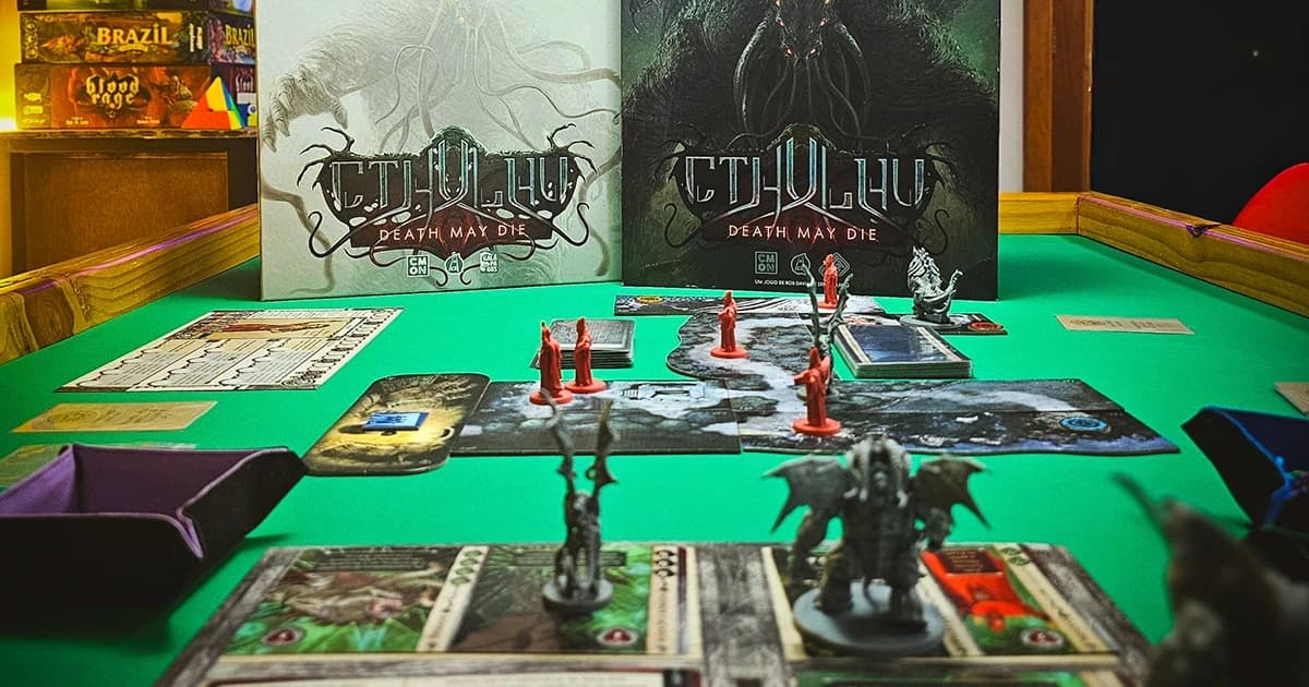 Partida do jogo Cthulhu: Death May Die montada numa mesa de RPG verde, com as duas caixas ao fundo, tabuleiros modulares, cartas e miniaturas.