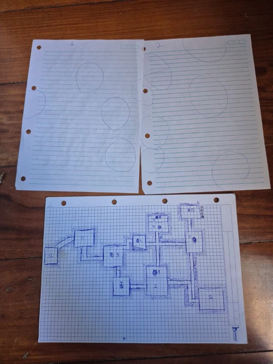 Foto de três folhas sobre um piso de madeira: duas com círculos a caneta e uma de papel quadriculado com um mapa de masmorra desenhado à mão em azul, com salas numeradas.
