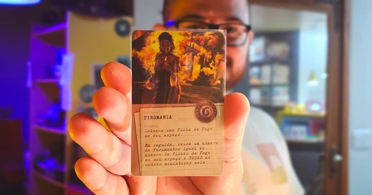 Mão segurando a carta de patologia Piromania do jogo Cthulhu: Death May Die, com um jogador desfocado ao fundo.