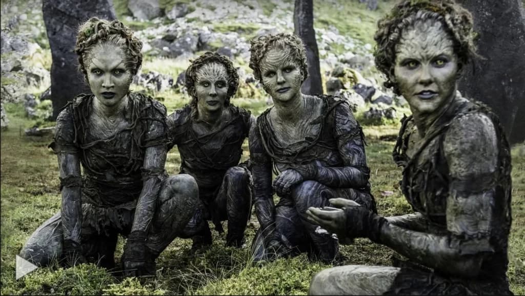 Quatro figuras de pele acinzentada e olhos claros agachadas na floresta, ao estilo dos Filhos da Floresta.