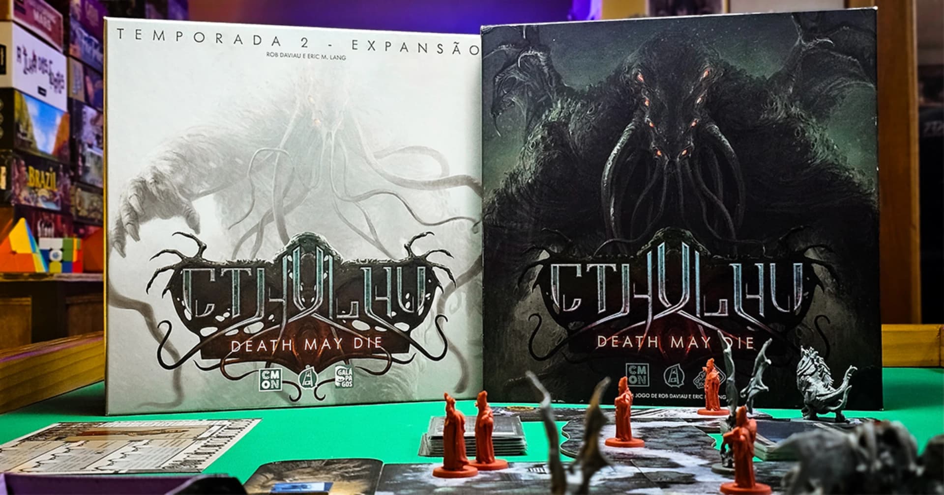 Duas caixas do jogo de tabuleiro Cthulhu: Death May Die (a expansão Temporada 2 e o jogo base) sobre uma mesa, com miniaturas de cultistas vermelhos à frente.